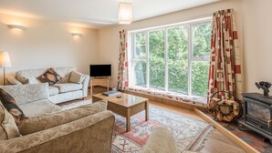 Ein gemütliches Wohnzimmer mit einer Couch, einem Couchtisch und einem Fernseher. Ein großes Fenster mit Vorhängen bietet Blick auf Bäume.