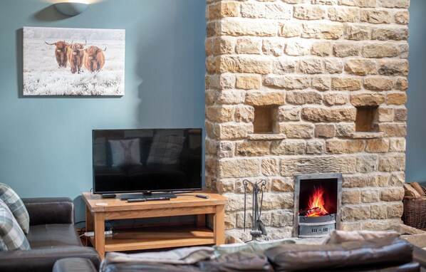 A living room with a stone fireplace, a flat-screen TV on a wooden stand, and a painting of Highland cattle on the wall. - The Old Cart House (York)