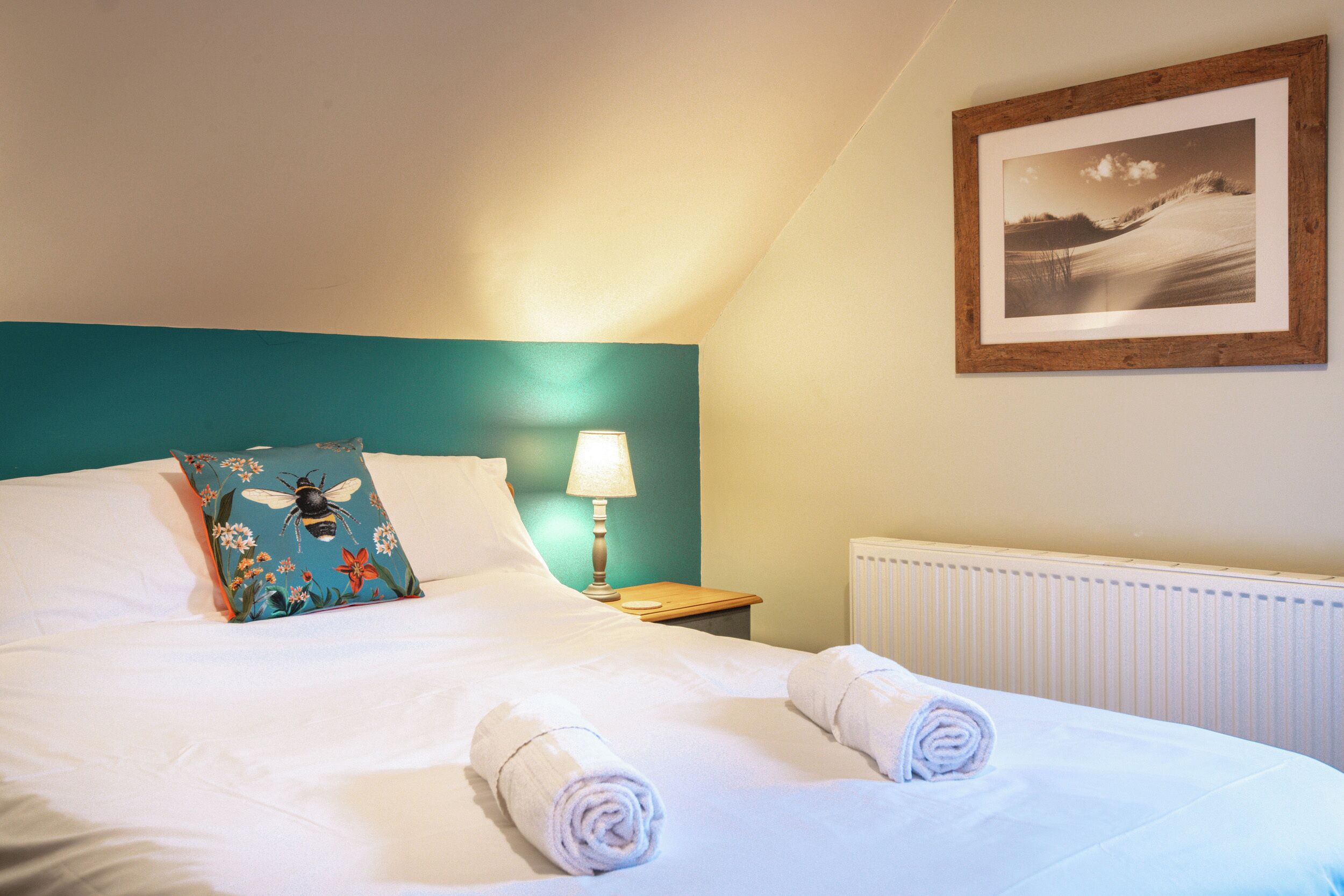 A neatly made bed with white linens and two rolled towels, a teal-colored pillow with a floral design, a bedside lamp, and a framed picture on the wall.