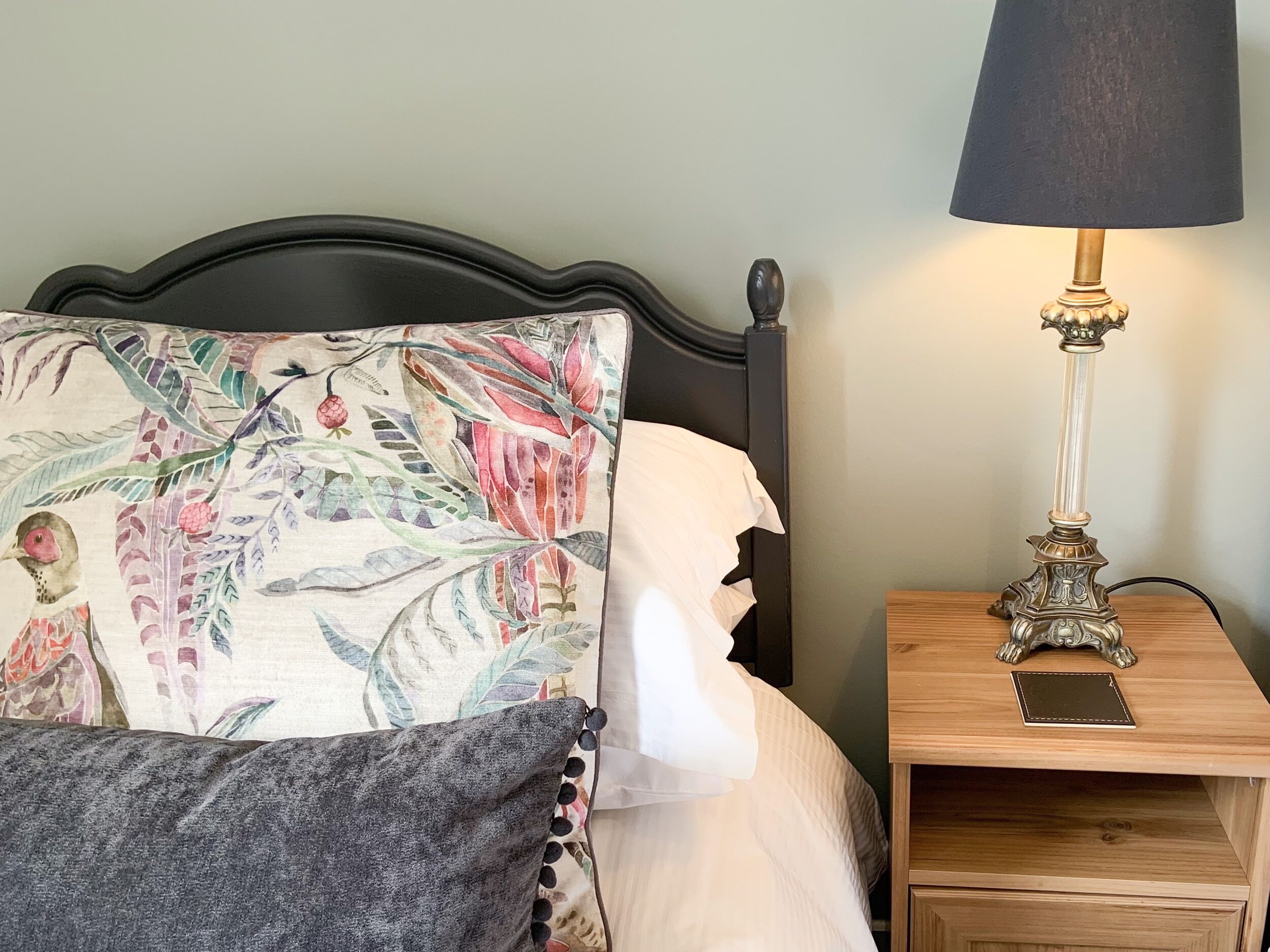 A bed with a floral pillow, a white pillow, and a gray throw blanket. There is a bedside table with a lamp and a smartphone on it.
