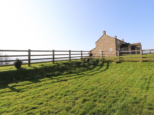 A stone house with a wooden fence in front, a grassy field, and a body of water in the background.