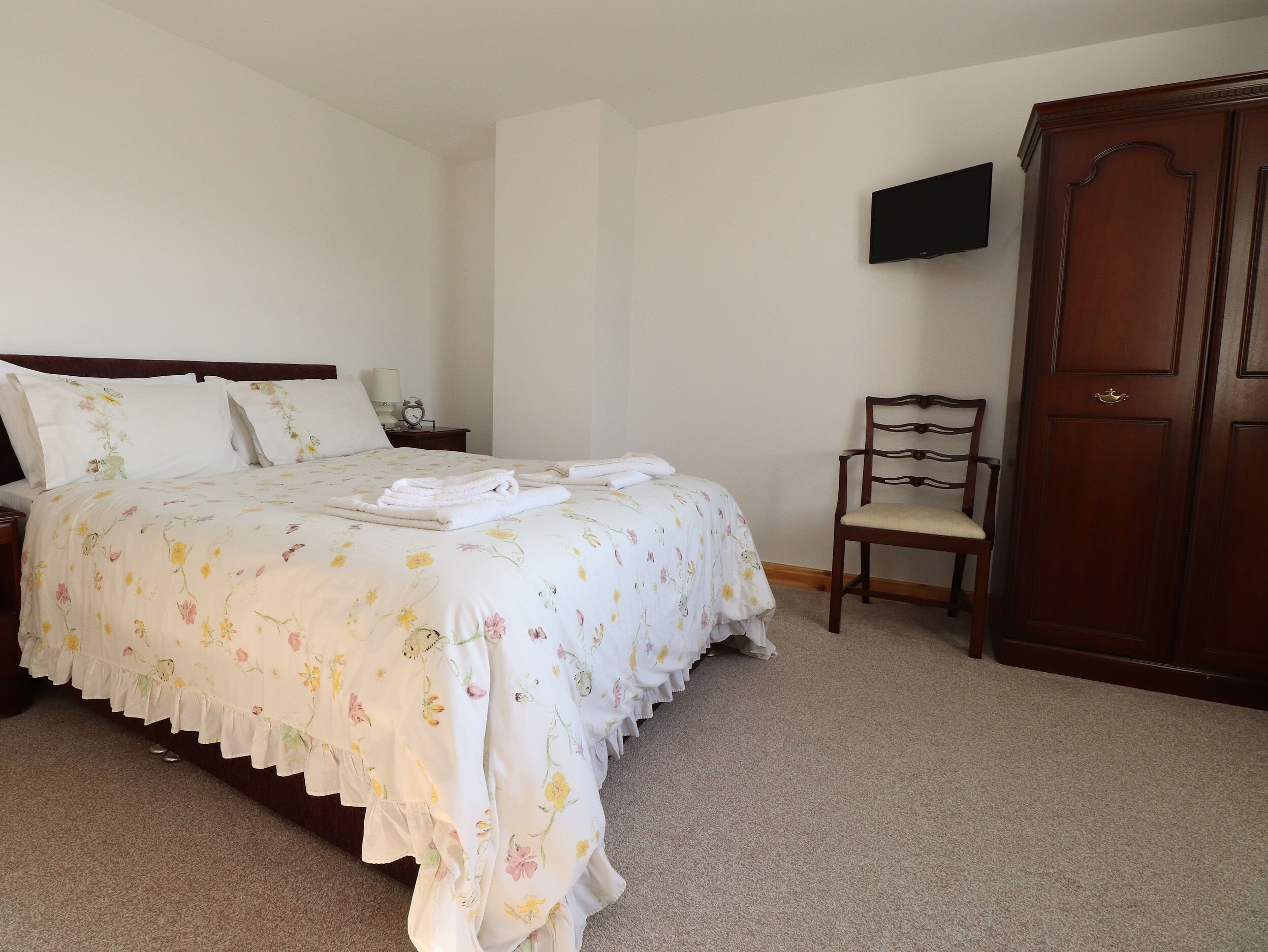 Una cama bien hecha con ropa de cama floral, una silla de madera y un armario en una habitación sencilla y despejada.