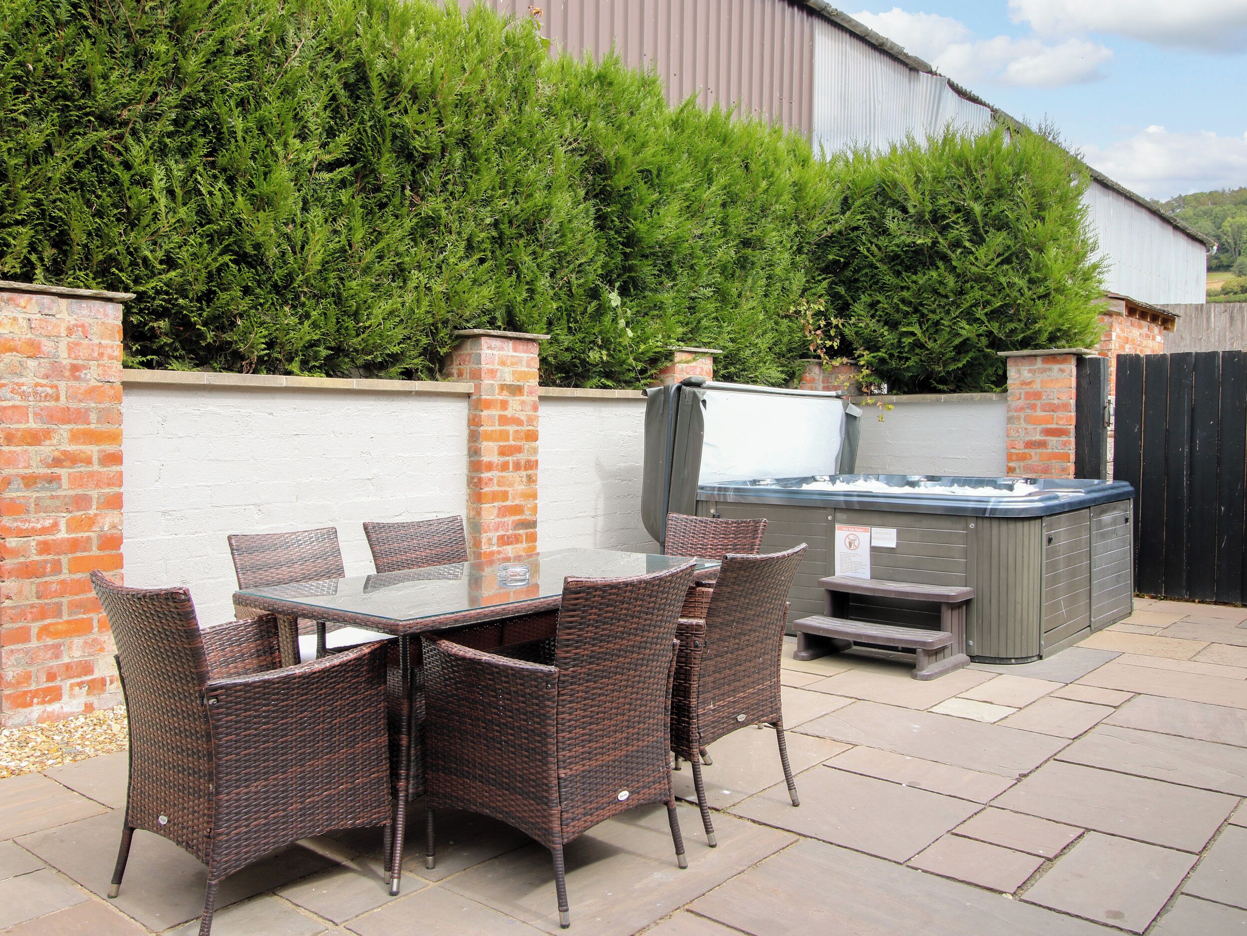 An outdoor patio with a glass-top table and wicker chairs, a hot tub, and a brick wall with greenery.