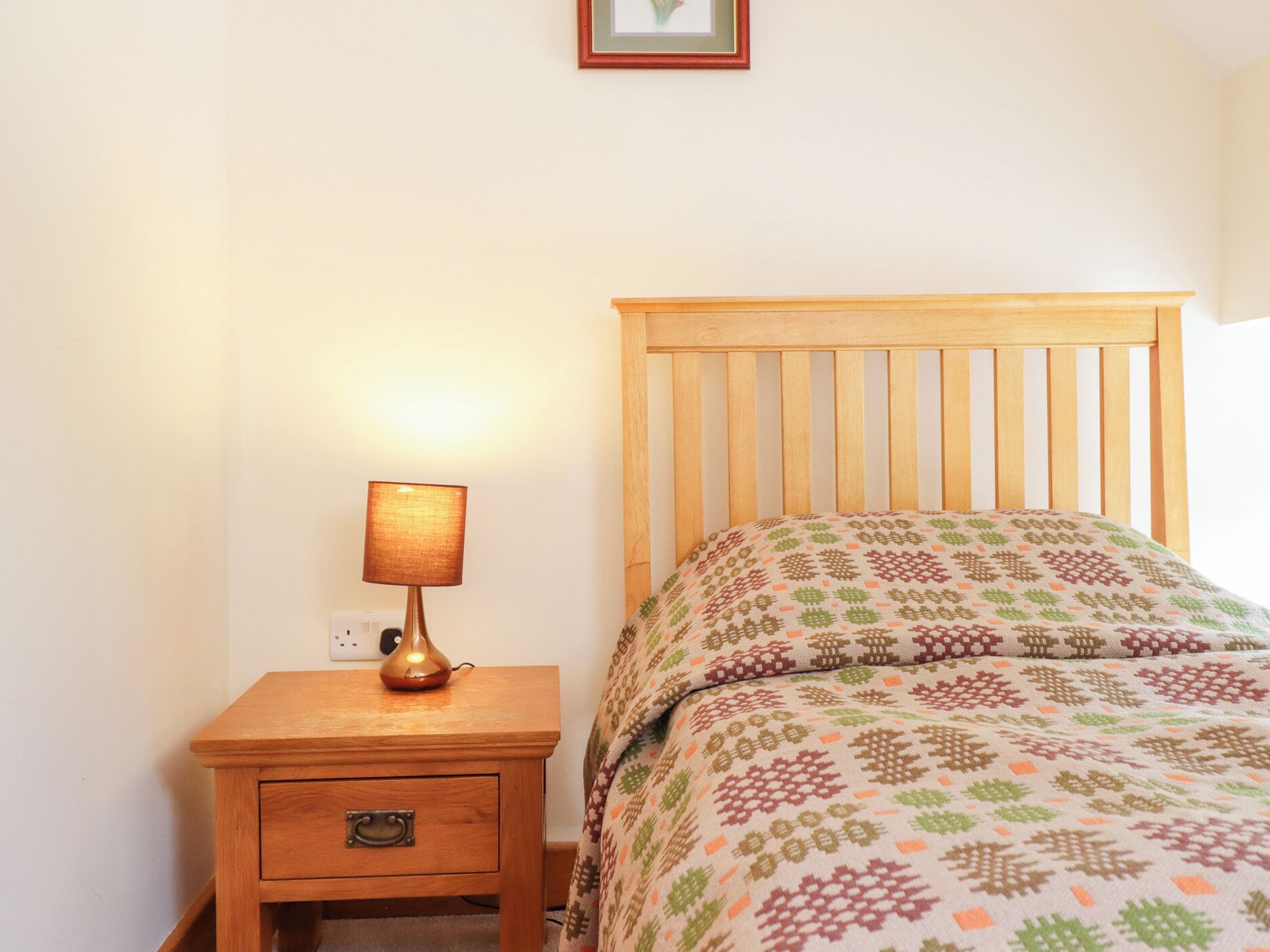 A bedroom with a wooden nightstand, a lamp, a patterned bedspread, and a framed picture on the wall.