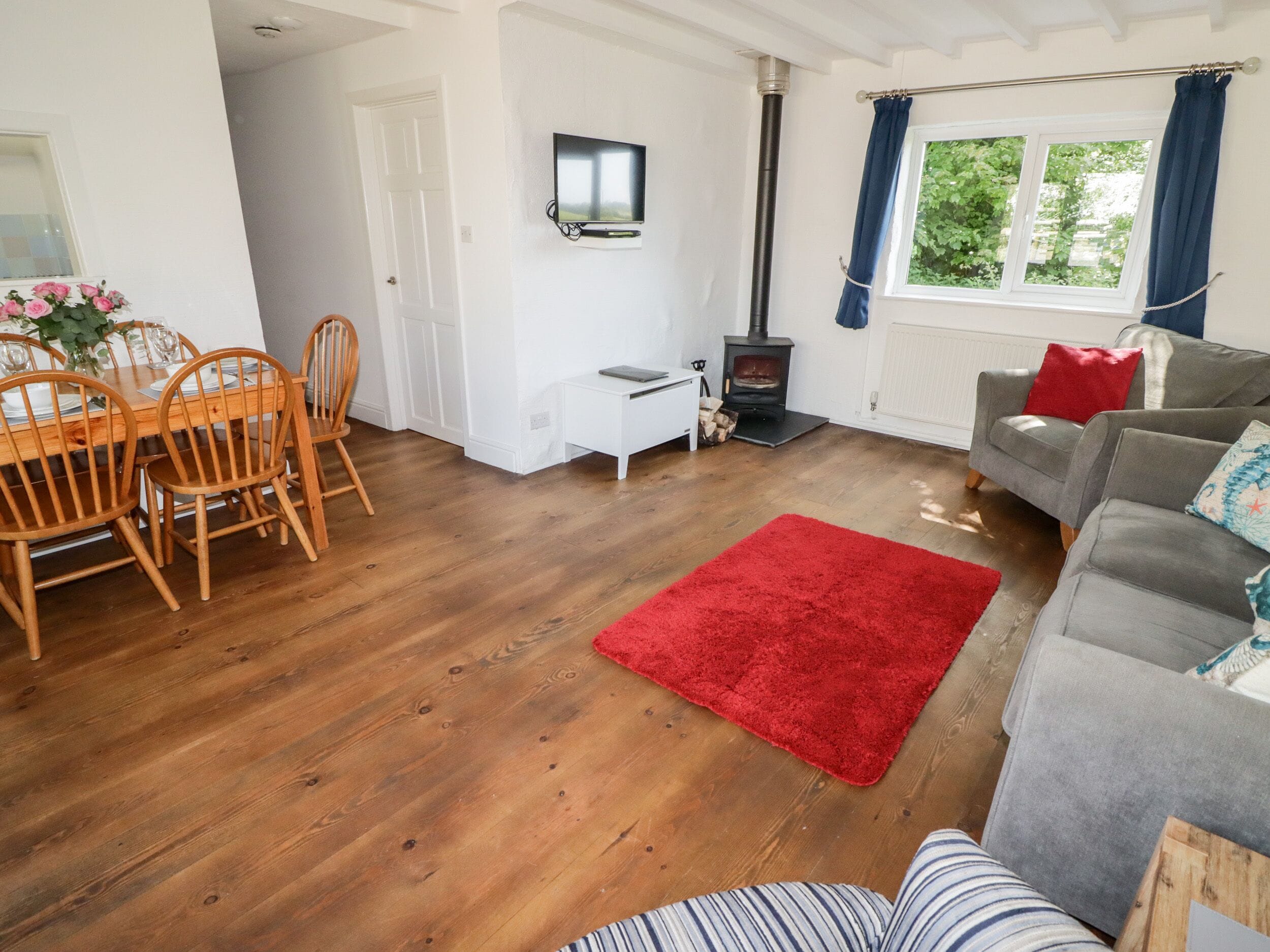 A living room with a wooden floor, a red rug, a grey sofa, and a dining area with wooden chairs and a table.