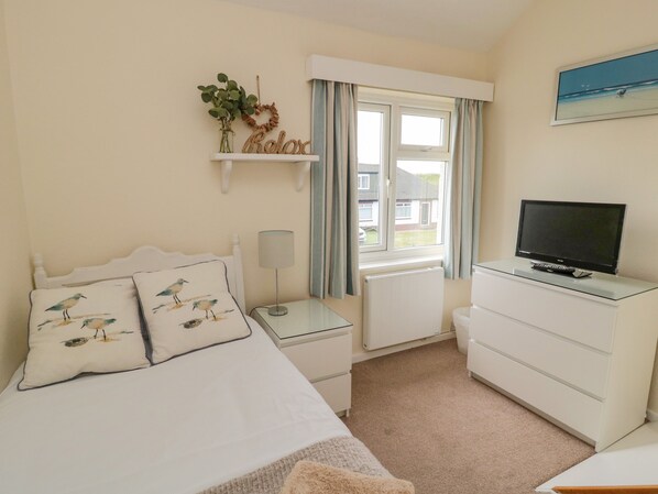 A bedroom with a bed, a TV, a nightstand, a dresser, a window with curtains, and a wall decoration. - Dune View (Chathill)