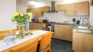 A modern kitchen with wooden cabinets, a dining table with chairs, and a vase of yellow flowers. - Penrallt Cottage (Y Felinheli)