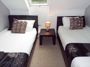 Ein Hotelzimmer mit zwei Betten, jedes mit einem dunklen Kopfteil und einem weißen Kissen. Dazwischen steht ein kleiner Tisch mit einer Lampe, darüber befindet sich ein Fenster.