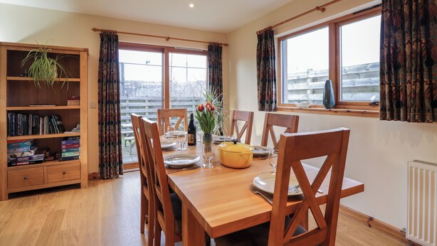 A dining area with a wooden table set for four, surrounded by chairs. There is a bookshelf to the left and a window with curtains on the right.