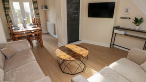 Een moderne woonkamer met een houten eettafel, beige banken, een televisie en een schoolbord met een welkomstboodschap.