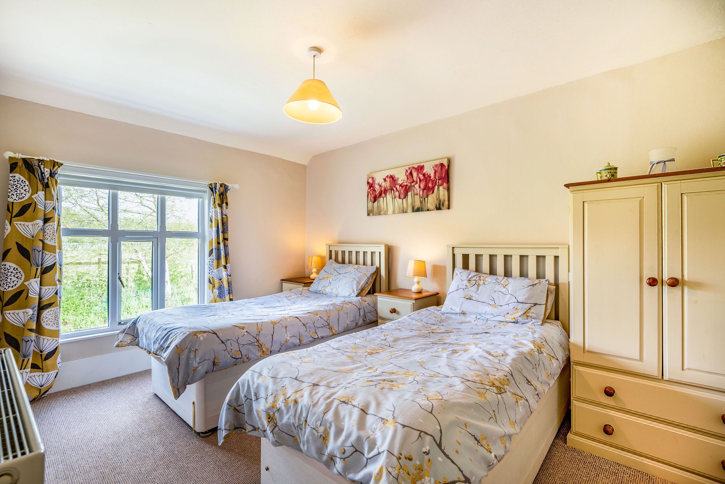 A bedroom with two beds, a window with curtains, a painting on the wall, and a cabinet.