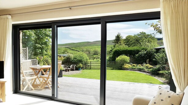 Une pièce spacieuse dotée d’une grande porte-fenêtre coulissante qui donne sur un patio extérieur. Le patio est agrémenté d’une table et de chaises en bois, entourés d ’une végétation luxuriante et d’un mur en pierre.