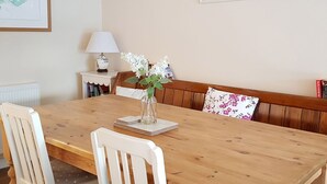 Un coin repas avec une table en bois, des chaises blanches, un vase avec des fleurs, un banc agrémenté d’un coussin à motifs floraux et deux tableaux encadrés accrochés au mur.