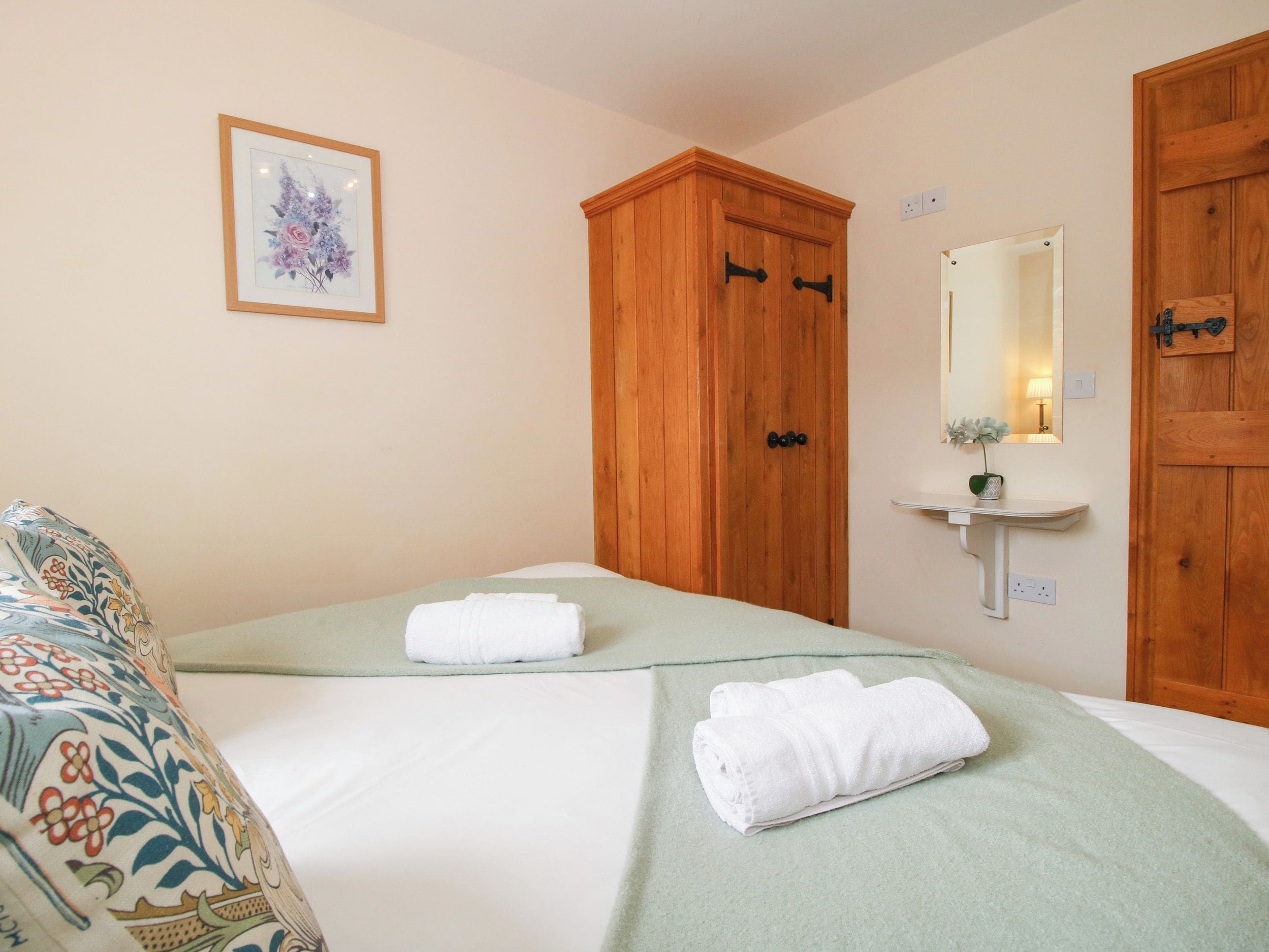 A hotel room with two beds, a wooden wardrobe, a bedside table with a lamp, and a framed floral artwork on the wall.