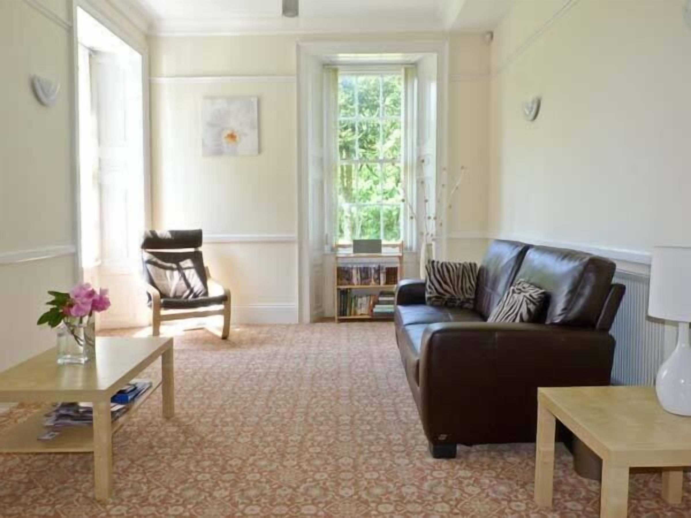 Un salon avec un canapé en cuir marron, une chaise en bois, une petite table sur laquelle est posé un vase de fleurs et une bibliothèque.