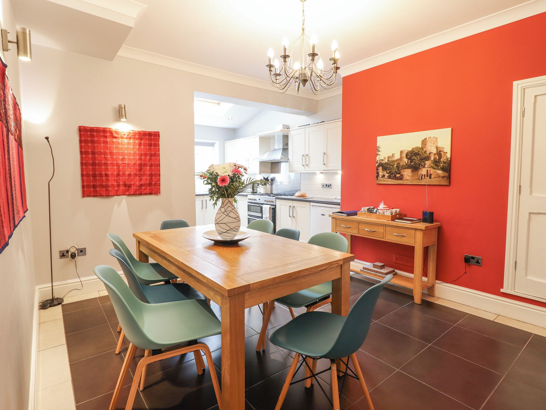 A dining area with a wooden table, teal chairs, a vase with flowers, and a red accent wall with a painting.
