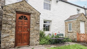 A stone building with a wooden door, a small table with chairs, and a grassy area in front. - Harber Scar (Settle)