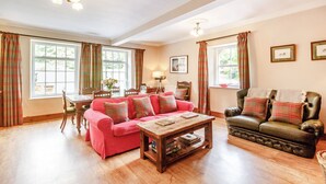 A cozy living room with a red sofa, a dark leather armchair, and a wooden coffee table. There are two windows with plaid curtains, a lamp on a side table, and framed pictures on the wall. - Reiver's Rest (Hexham)