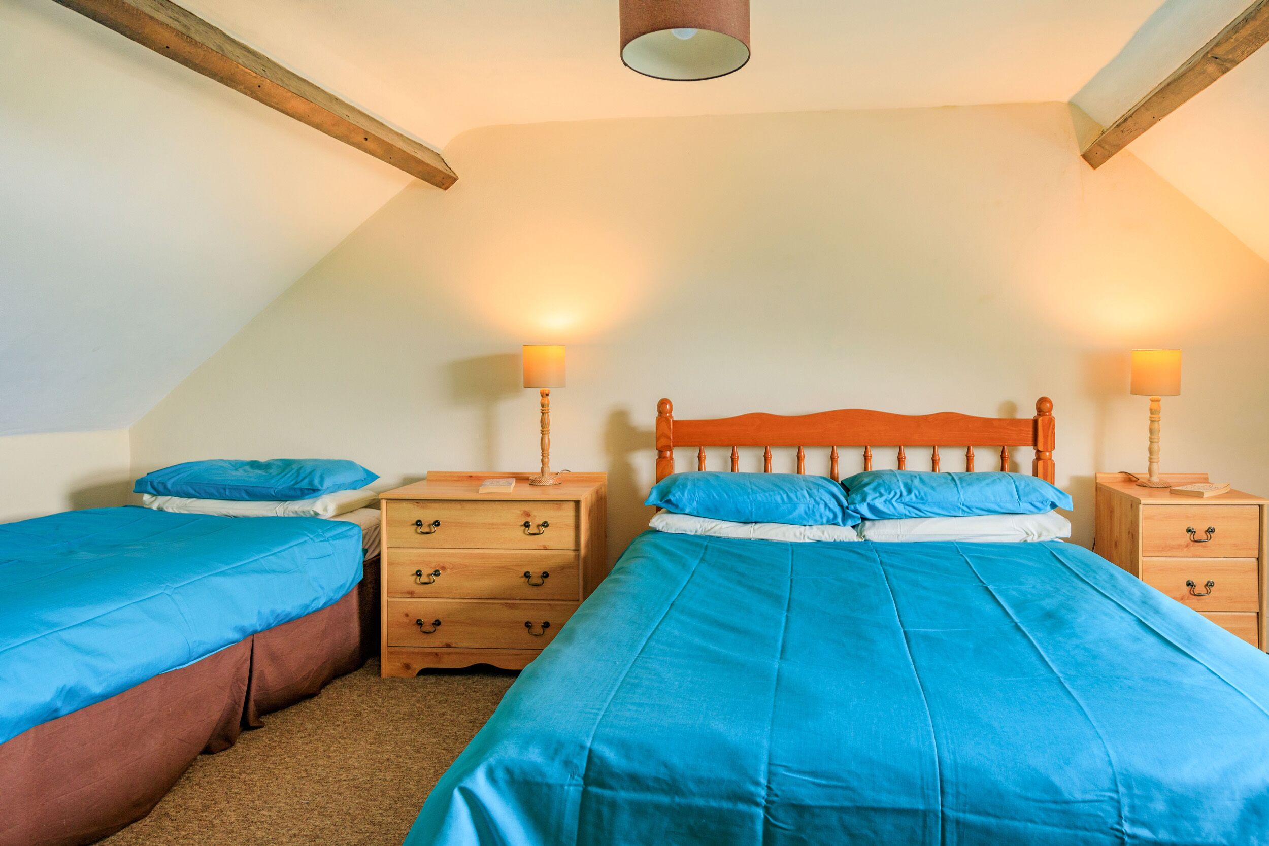 Un dormitorio con dos camas individuales, cabeceros de madera, mesitas de noche con lámparas y techo inclinado.