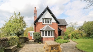 A traditional brick house with a stone patio, a wooden table, and a garden with various plants and a tree. - Gun End Cottage (Macclesfield)