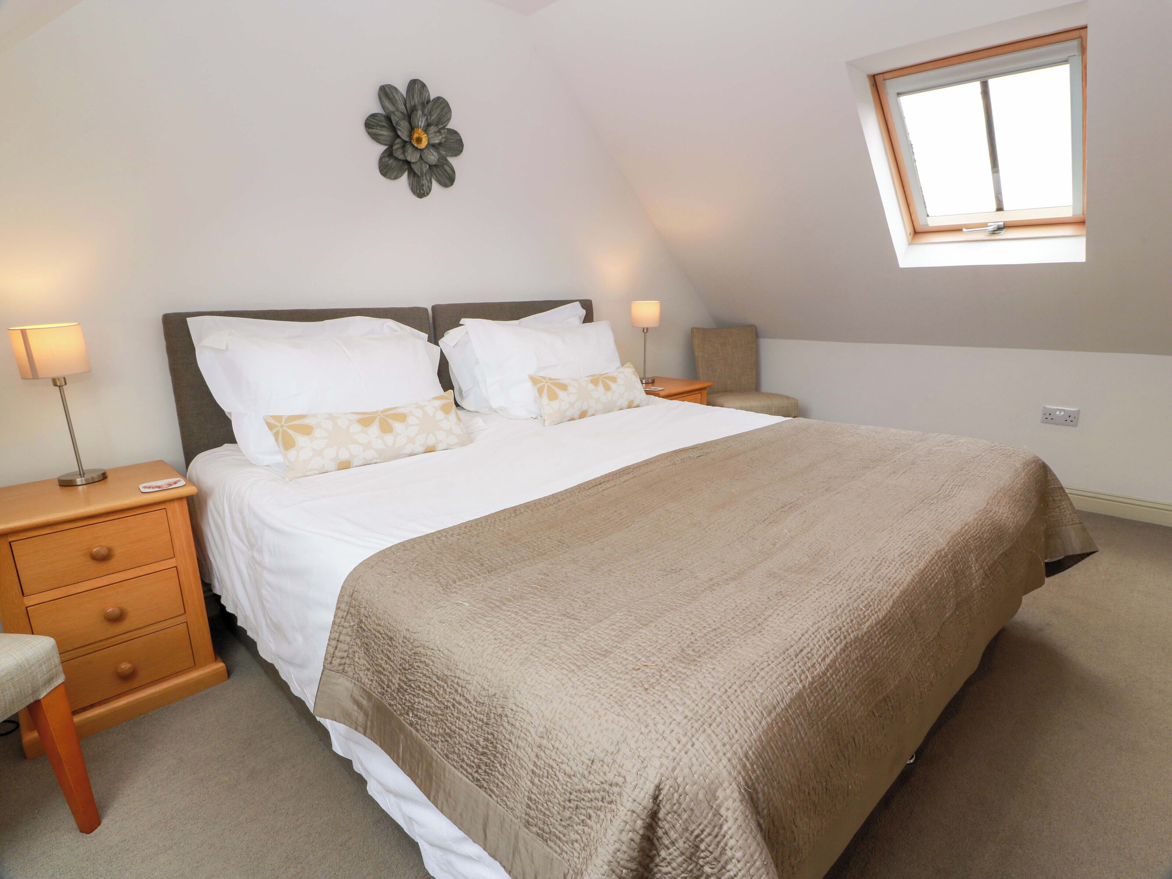 A bedroom with a large bed, two bedside tables, a nightstand, a lamp, a chair, a window, and a wall decoration.