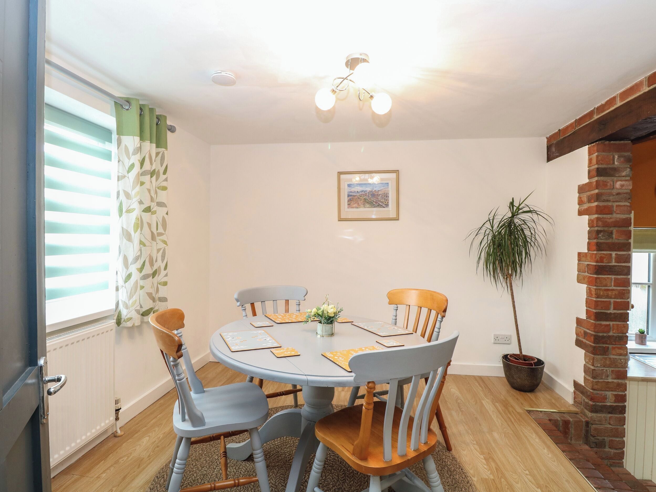 A dining area with a round table, four chairs, a potted plant, a framed picture on the wall, and a brick fireplace.