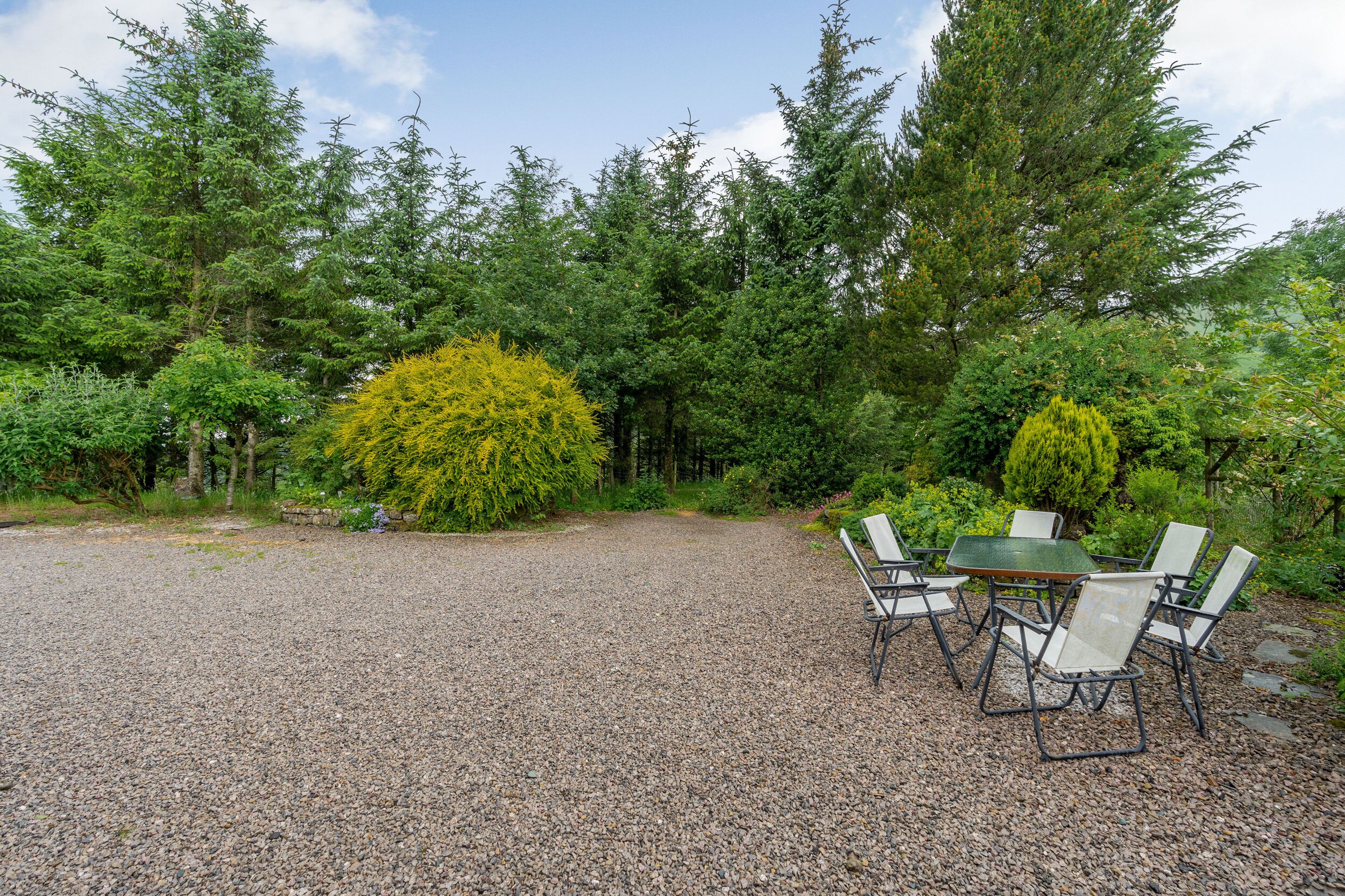 A gravel patio with a round table and four chairs, surrounded by dense greenery and tall trees.
