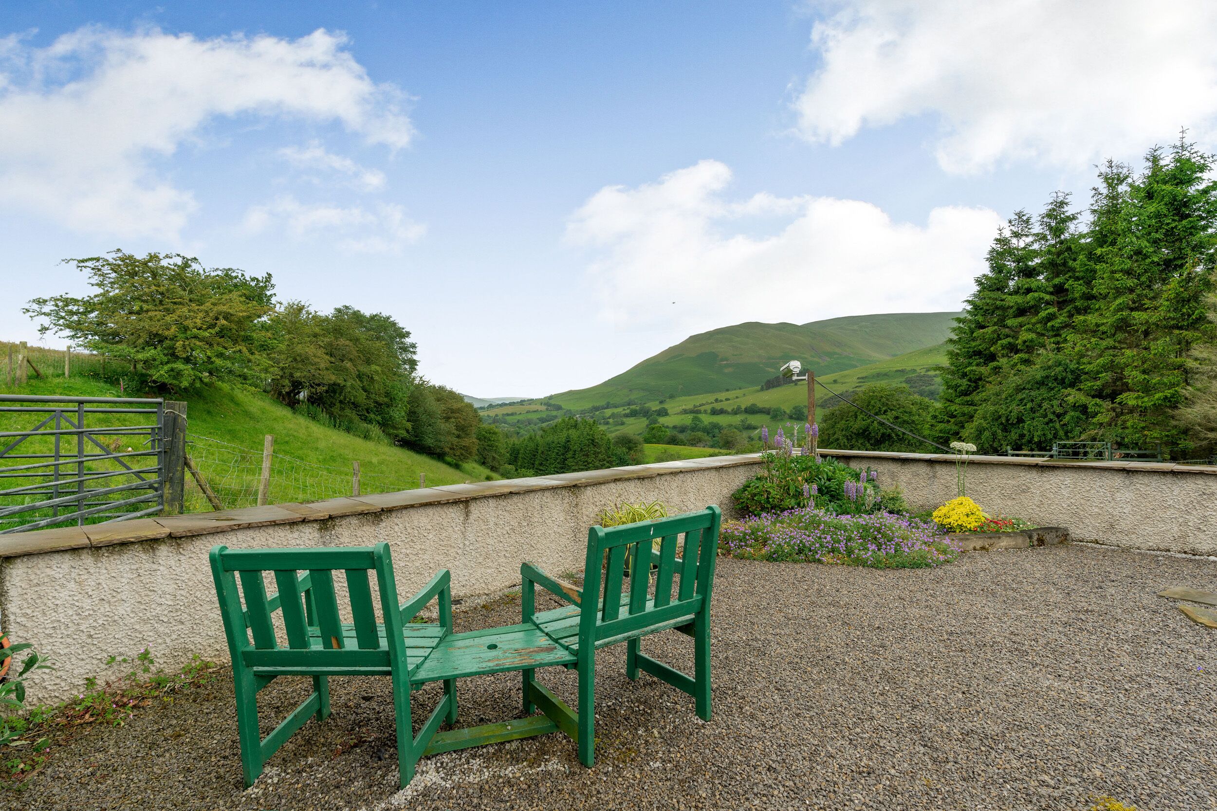 A green bench and table set on a gravel area with a view of green hills and a clear sky.