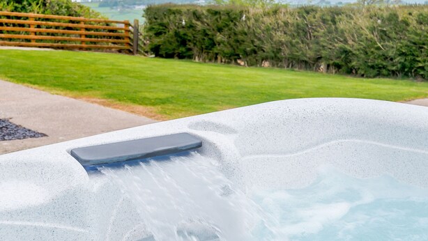 A hot tub with a waterfall feature, surrounded by a grassy area and a wooden fence. - Pen Y Gaer (Denbigh)