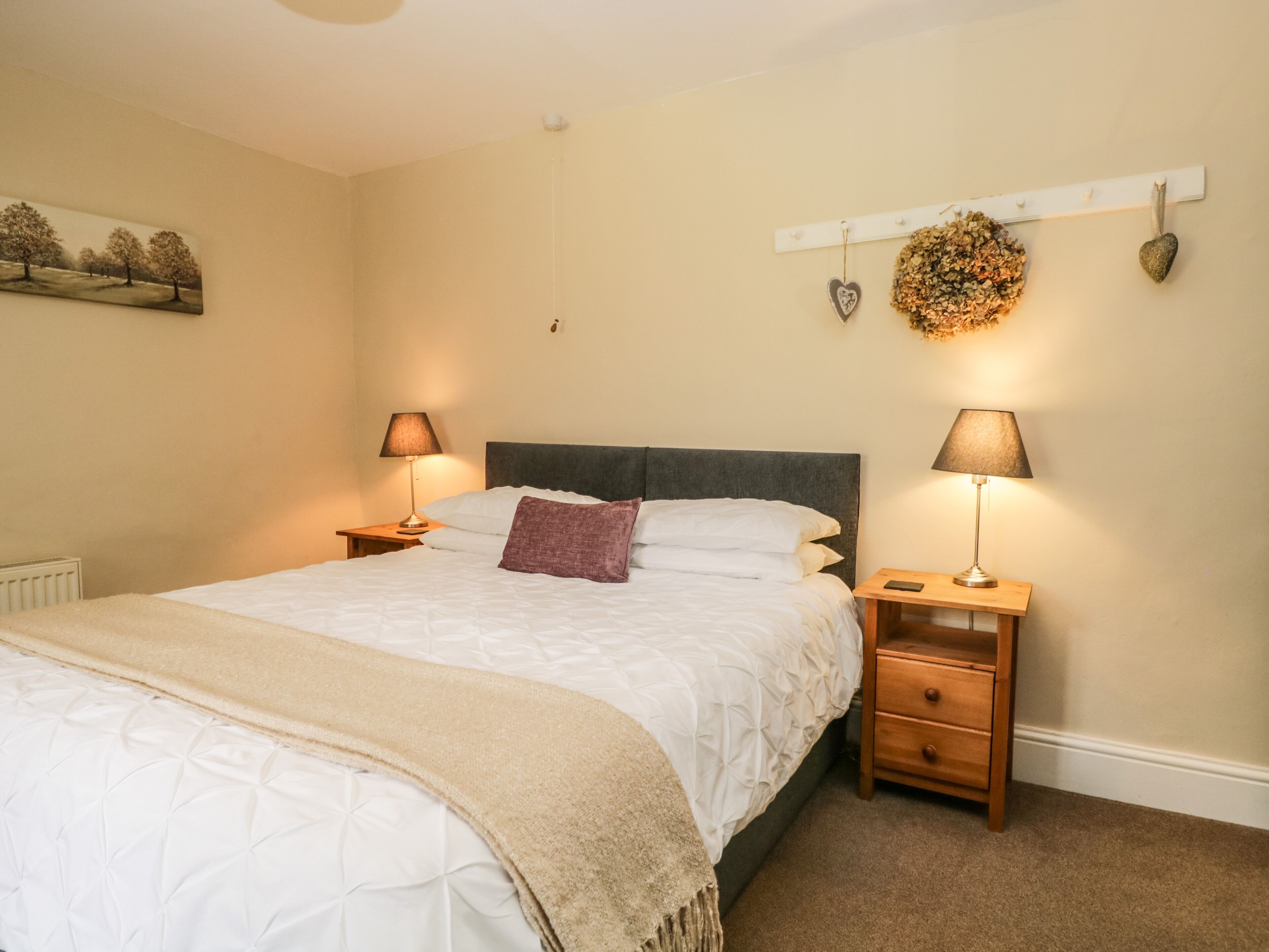 Una cama bien hecha con un edredón color beige, dos mesitas de noche con lámparas, un tocador de madera, un estante de pared con objetos decorativos y un cuadro en la pared.
