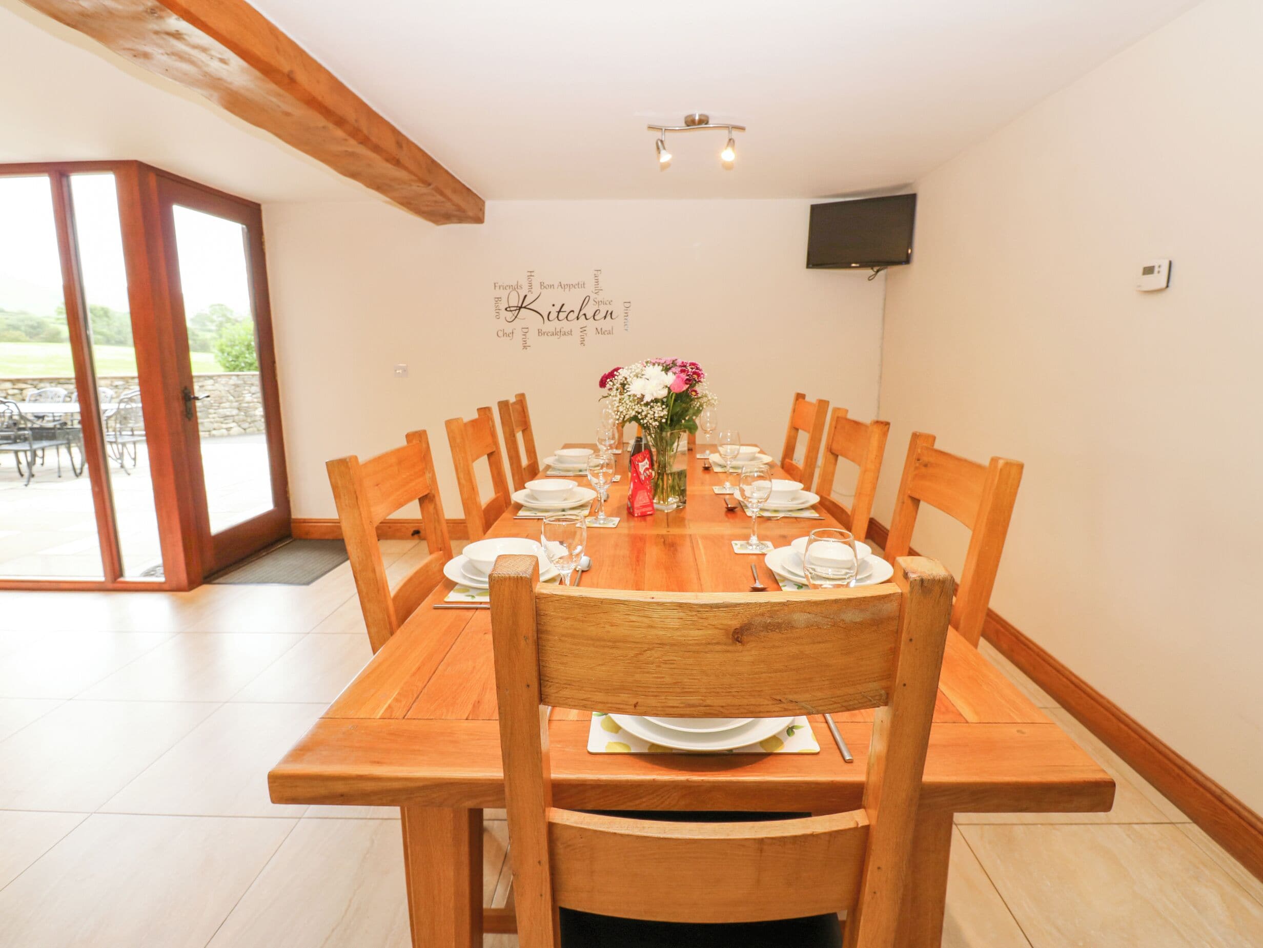 A dining area with a wooden table set for six, surrounded by wooden chairs. A word cloud on the wall behind the table includes words like "Chef," "Breakfast," and "Wine."