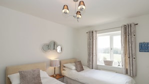 A bedroom with two beds, a bedside table with a lamp, a window with curtains, and a wall-mounted mirror. - Waddleduck (Whitby)