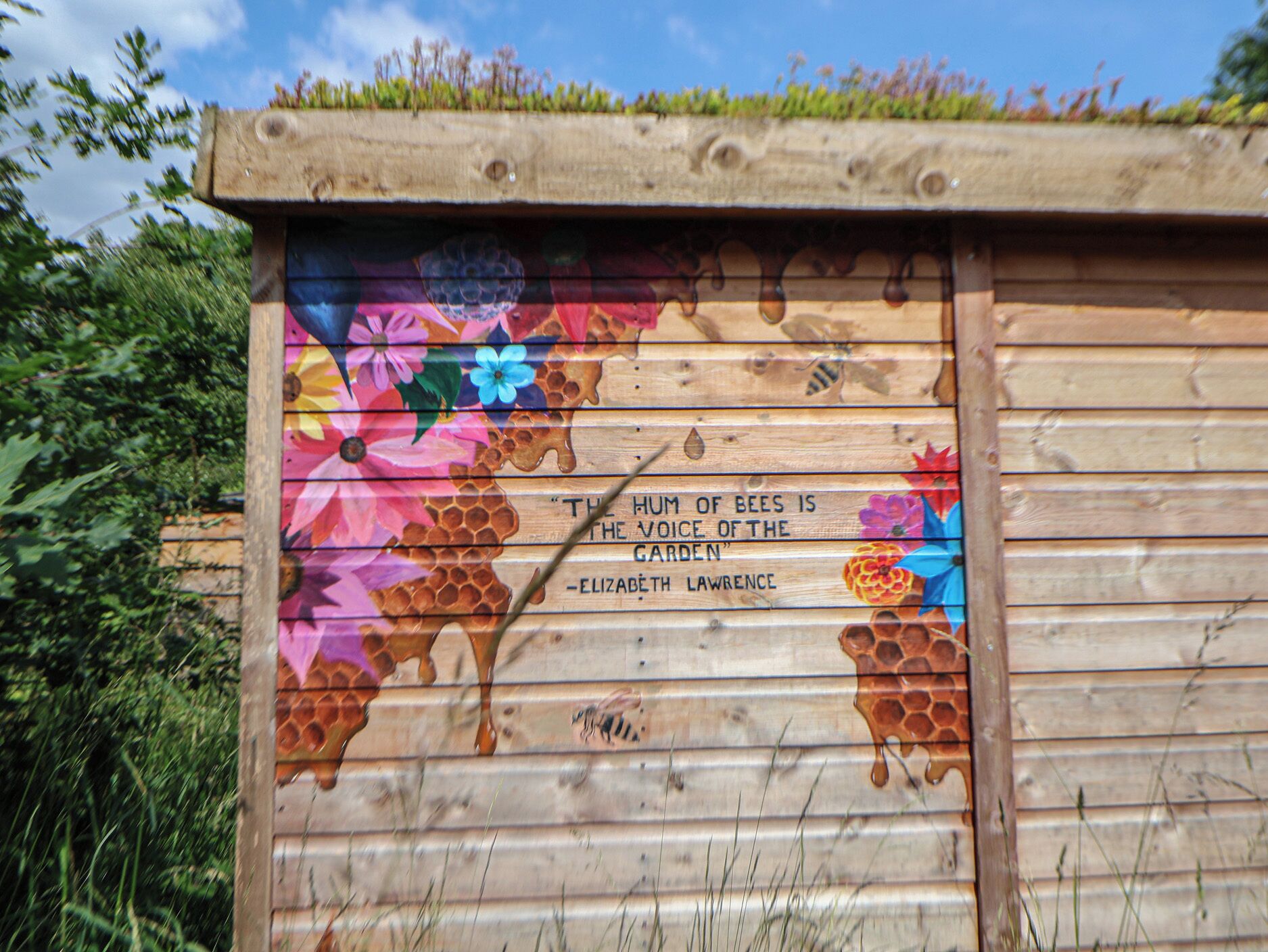Une structure en bois ornée d’une fresque représentant des abeilles et des fleurs, ainsi que d’une citation d’Elizabeth Lawrence.