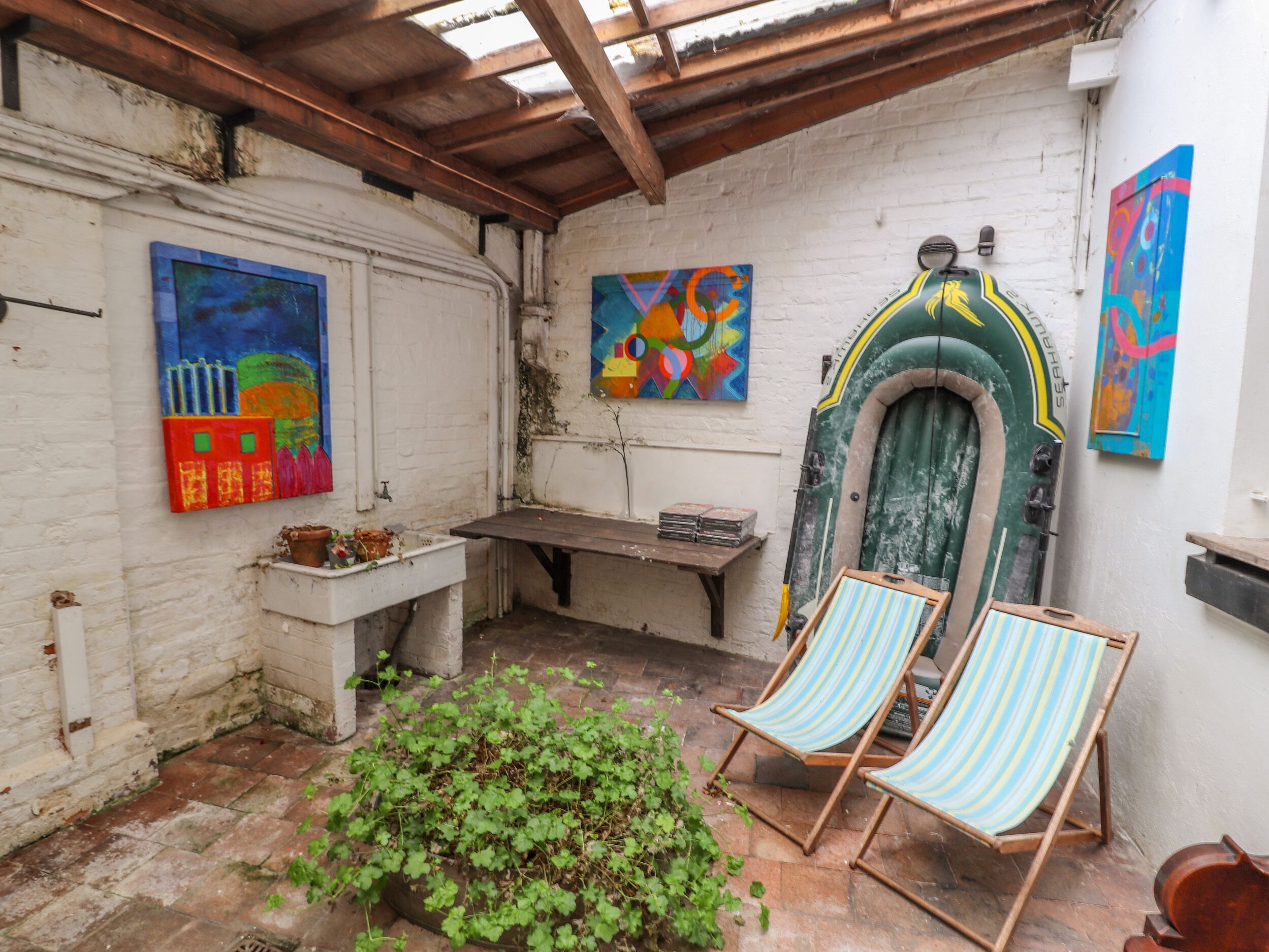 A rustic courtyard with a wooden bench, two striped lounge chairs, and colorful abstract paintings on the walls.