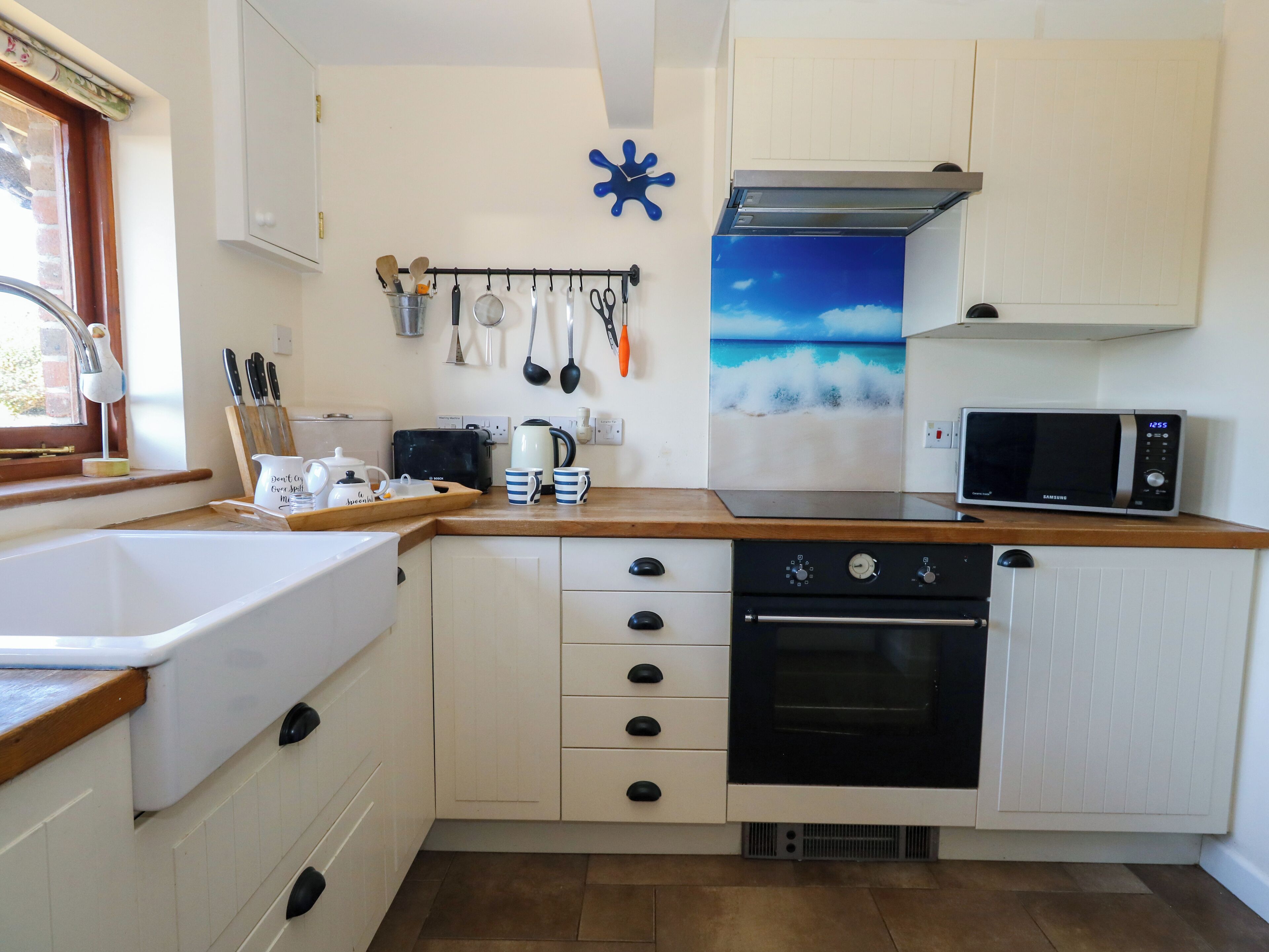 A compact kitchen with a microwave, toaster, and kettle. There is a window with a curtain, and a wall-mounted rack holding kitchen utensils.