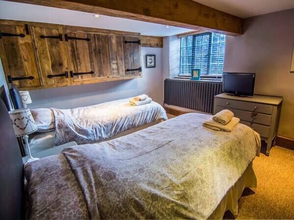 A bedroom with two beds, wooden cabinets, a radiator, a television, and a window with blinds. - The Malthouse (Matlock)