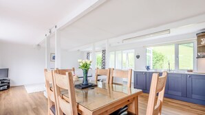 Un comedor espacioso con una mesa y sillas de madera, un florero con flores, y una cocina al fondo con armarios azules y una ventana.