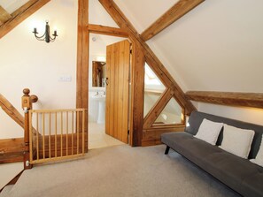A loft bedroom with exposed wooden beams, a wooden staircase, a grey sofa, and a white bathroom visible through an open door. - Trooper's Barn (Craven Arms)