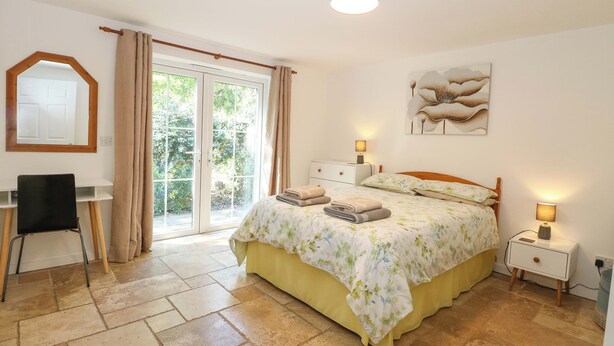 A bedroom with a bed, a desk, a chair, a nightstand, a lamp, a mirror, and a view through a glass door. - Mill Lodge, Brisley Accessible Norfolk Cottage Sleeping 6, Dog-friendly (Dereham)