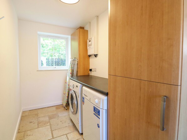 A compact kitchen with a washing machine, wooden cabinets, a window, and a door with a handle. - Mill Lodge, Brisley Accessible Norfolk Cottage Sleeping 6, Dog-friendly (Dereham)