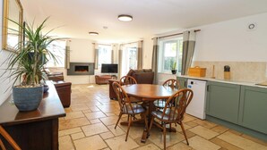 A dining area with a table and chairs, a fireplace, a television, and a kitchen area with cabinets and a window. - Mill Lodge, Brisley Accessible Norfolk Cottage Sleeping 6, Dog-friendly (Dereham)