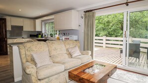 Un salon avec un canapé, une table basse avec un plateau en verre, une cuisine en arrière-plan et un balcon donnant sur un espace verdoyant.