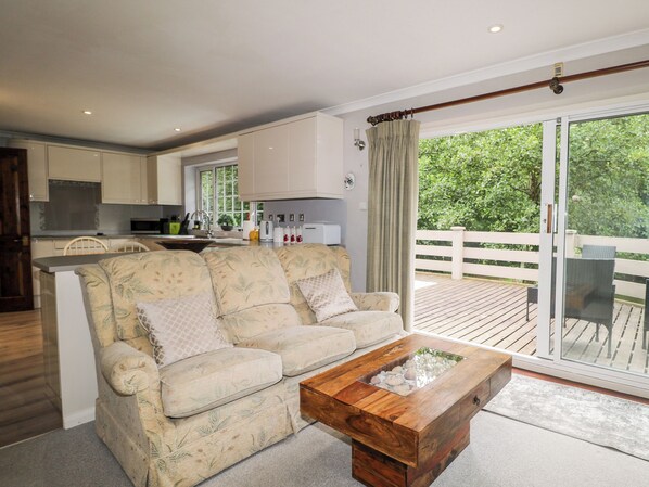 A living room with a sofa, a coffee table with a glass top, a kitchen in the background, and a balcony with a view of greenery. - Miswells Cottages - Lake View (Crawley)