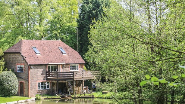Une maison avec un toit en pente, une terrasse en bois et un étang à l’avant.