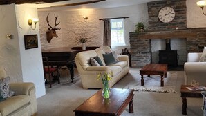A cozy living room with a stone fireplace, a sofa, a coffee table, and a dining area with a piano. - High Kiln Bank Cottage (Broughton in Furness)