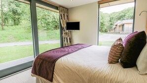 Un dormitorio con una cama grande, un televisor montado en la pared y vistas a un paisaje verde a través de amplias ventanas.