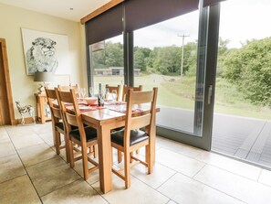 A dining area with a table set for four, wooden chairs, and a view of a green landscape through large windows. - Neddy Cut (Ulverston)