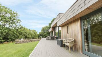 A modern outdoor deck with a stone wall, a wooden bench, and a glass door leading to a covered area.