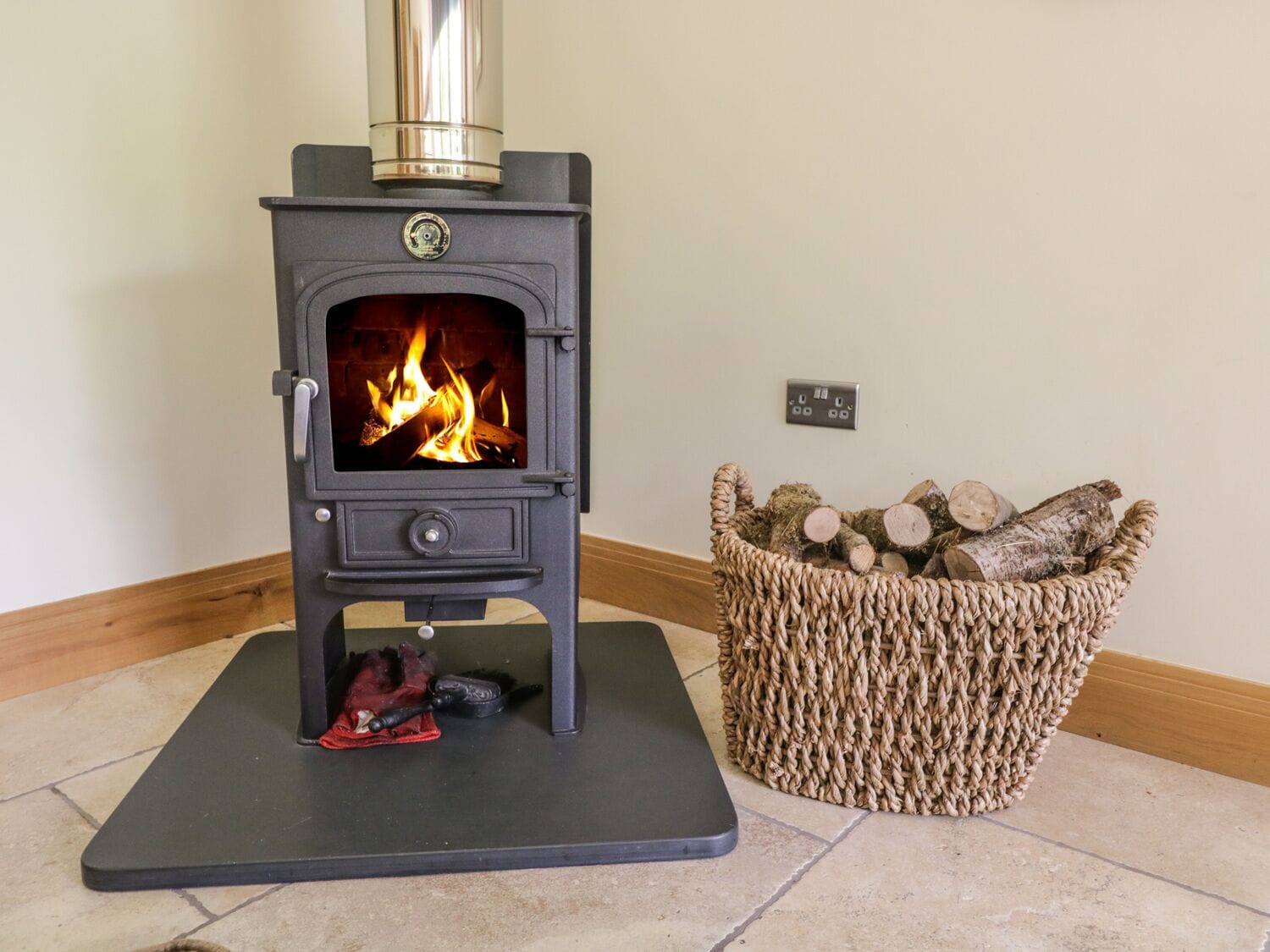 A black wood-burning stove with a glass door, a red cloth, and a woven basket of logs beside it.