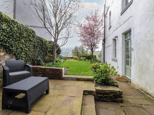 A patio with wicker furniture, a stone pathway, a hedge, and a view of a garden with a tree and a building in the background.